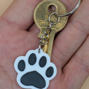May include: A white and black paw print keychain with a gold key and silver key ring. The paw print design has a black paw pad and black toe beans. The keychain is held in a hand.
