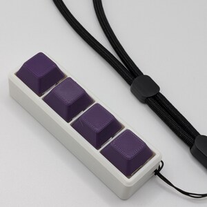 May include: A white rectangular case holds four purple keycaps. The keycaps have a textured surface and are connected to a black lanyard. The lanyard has a black plastic clasp.