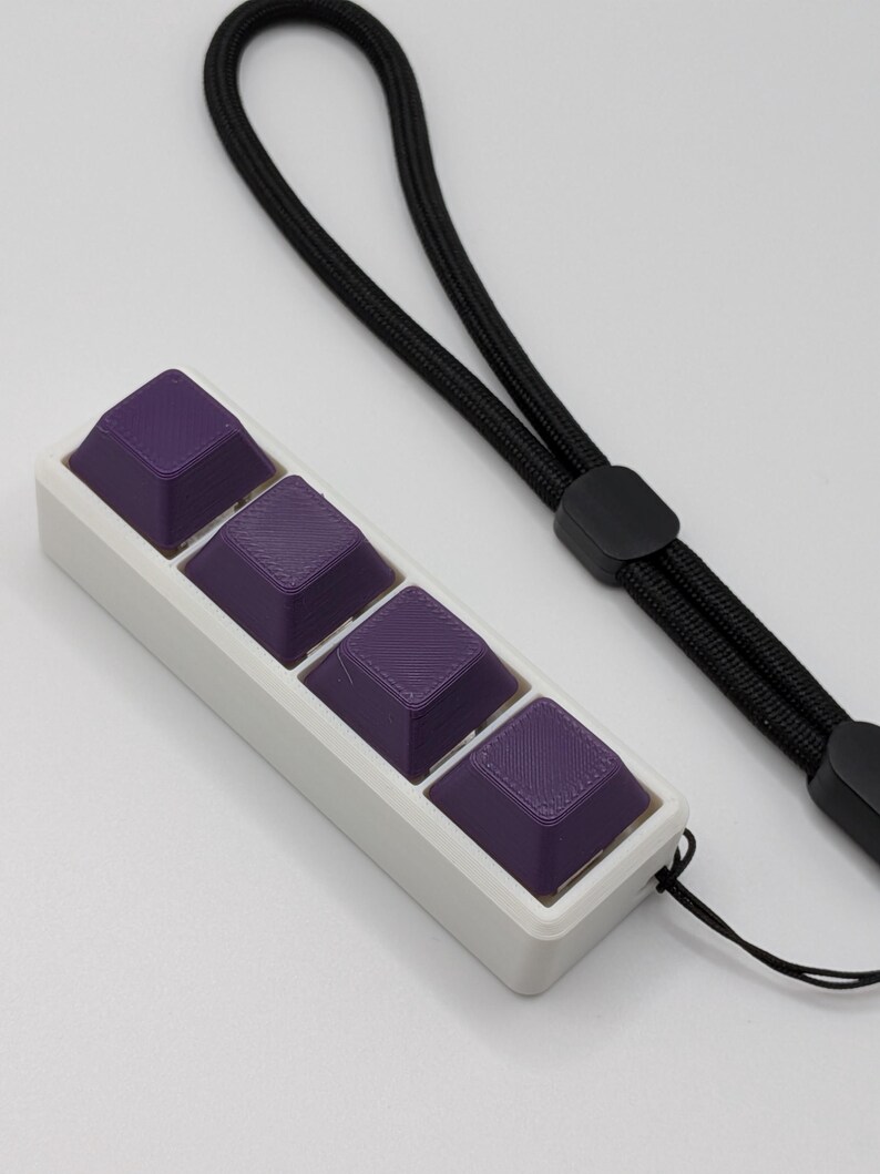 May include: A white rectangular case holds four purple keycaps. The keycaps have a textured surface and are connected to a black lanyard. The lanyard has a black plastic clasp.