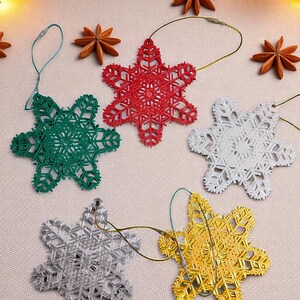 May include: Five snowflake-shaped Christmas ornaments in red, green, white, silver, and gold. Each ornament has a detailed, openwork design and a loop for hanging. The ornaments are arranged on a light-colored surface, with star anise and greenery in the background.
