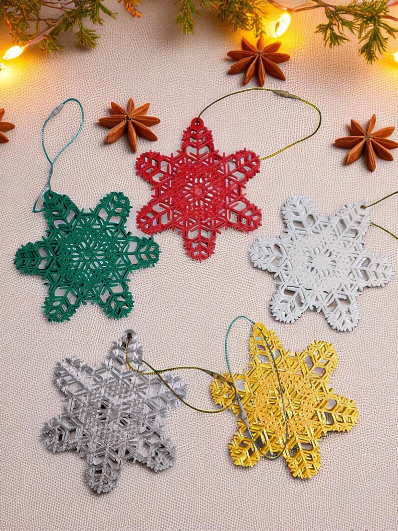 May include: Five snowflake-shaped Christmas ornaments in red, green, white, silver, and gold. Each ornament has a detailed, openwork design and a loop for hanging. The ornaments are arranged on a light-colored surface, with star anise and greenery in the background.
