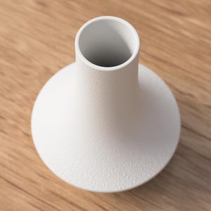 May include: A white ceramic vase with a textured surface. The vase has a rounded base and a cylindrical neck. The vase is sitting on a wooden surface.