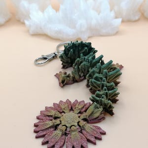 May include: A unique keychain featuring a detailed, abstract design. The keychain has a silver clasp and a decorative element in shades of green, brown, and purple. The design resembles a stylized plant or crystal formation.
