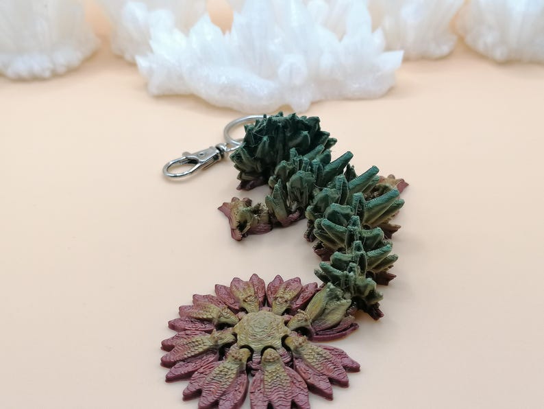 May include: A unique keychain featuring a detailed, abstract design. The keychain has a silver clasp and a decorative element in shades of green, brown, and purple. The design resembles a stylized plant or crystal formation.