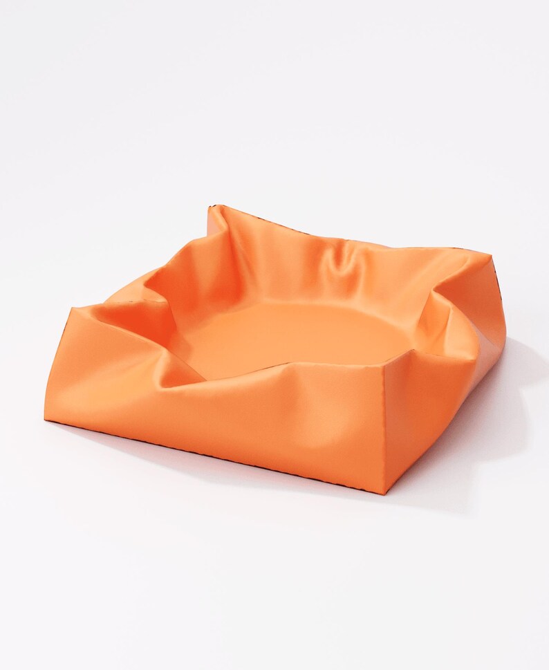 May include: An orange square tray with folded edges. The tray has a smooth, matte finish and a shallow interior. The corners are folded up, creating a unique, sculptural shape. The tray is set against a white background.