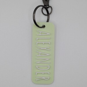 May include: A light green rectangular keychain with the name "ALEXANDER" in white lettering. The keychain has a dark gray metal clasp and ring for attaching to keys or a bag. The background is a solid light gray.
