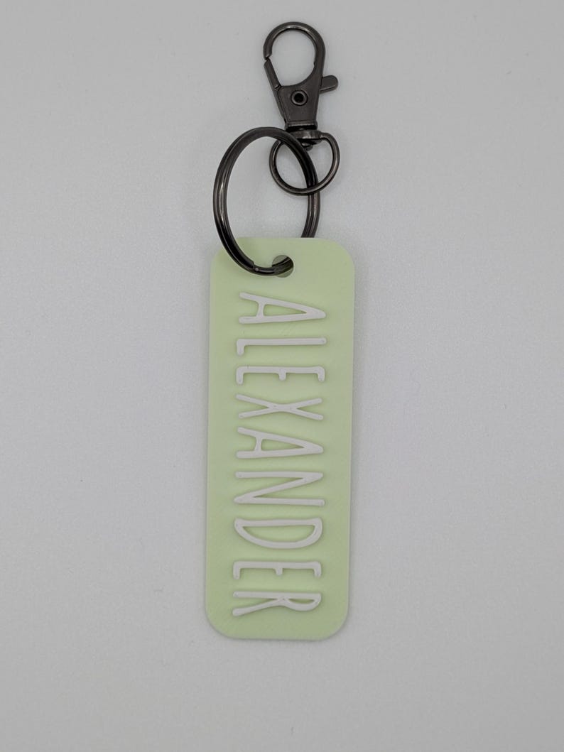 May include: A light green rectangular keychain with the name "ALEXANDER" in white lettering. The keychain has a dark gray metal clasp and ring for attaching to keys or a bag. The background is a solid light gray.