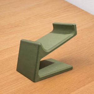 May include: A sage green, angled phone stand. The stand has a rectangular base, a vertical support, and a sloped top to hold a phone. The stand is made of a solid material and is designed to hold a phone at an angle.