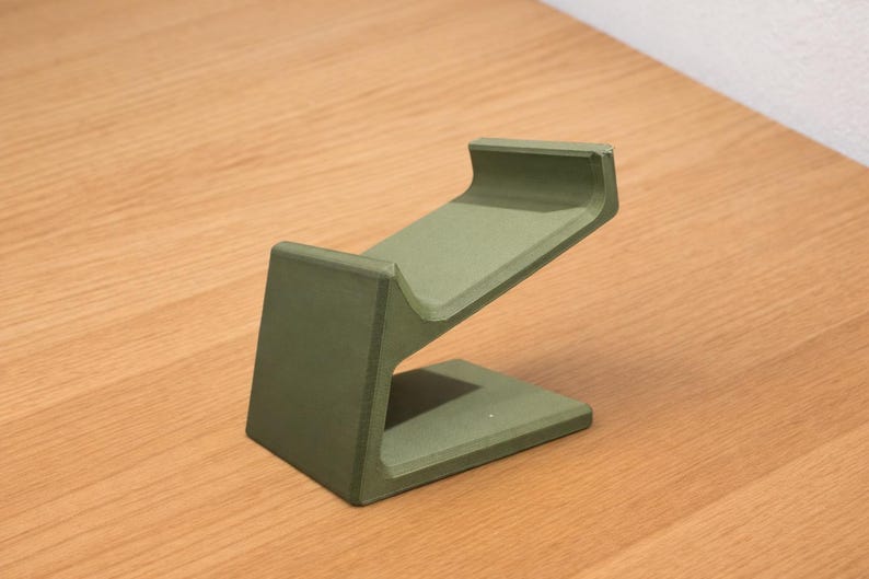 May include: A sage green, angled phone stand. The stand has a rectangular base, a vertical support, and a sloped top to hold a phone. The stand is made of a solid material and is designed to hold a phone at an angle.