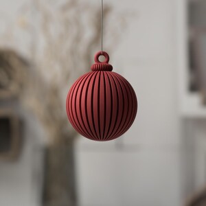 May include: A red, spherical ornament with vertical grooves and a loop for hanging. The decorative ball is suspended by a thin string, ideal for holiday or seasonal decor. The ornament's design features a ribbed texture.