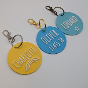 May include: Three personalized keychains in yellow, blue, and light blue. Each keychain is round with a name in white raised lettering. The yellow keychain has a rainbow graphic, the blue keychain has the text "CLASS 3B", and the light blue keychain has a star.