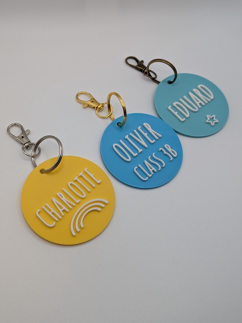 May include: Three personalized keychains in yellow, blue, and light blue. Each keychain is round with a name in white raised lettering. The yellow keychain has a rainbow graphic, the blue keychain has the text "CLASS 3B", and the light blue keychain has a star.