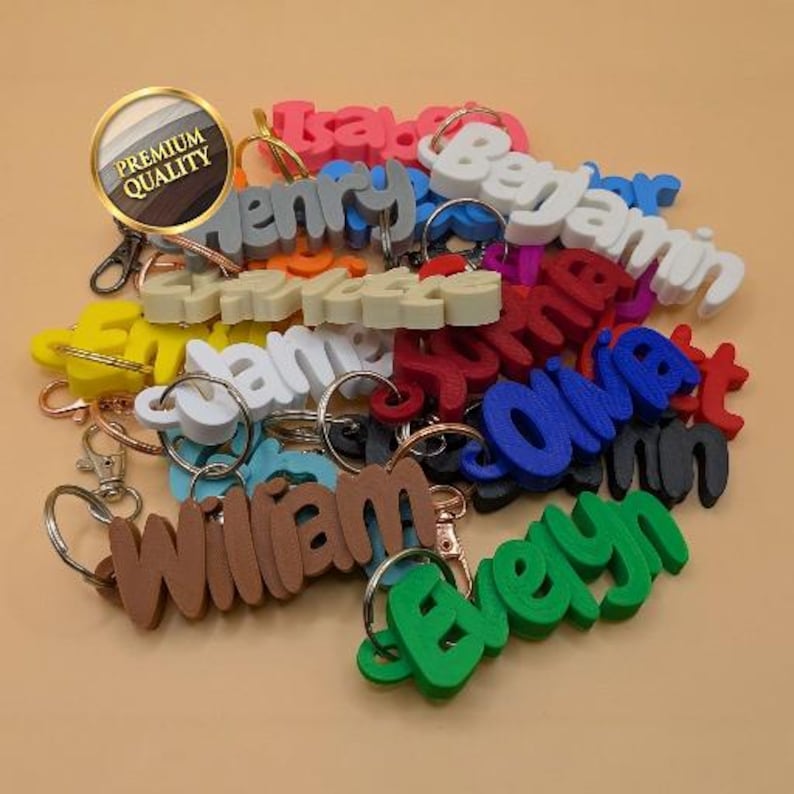 May include: A collection of 3D printed name keychains in various colors including red, blue, green, white, brown, and gray. Each keychain features a custom name in a bold, raised font attached to a metal split ring or lobster clasp hardware.