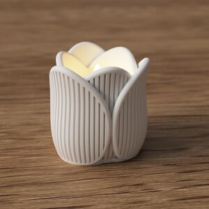 May include: A white, tulip-shaped candle holder with vertical ribbed detailing. The interior glows with a soft, warm light. The candle holder is made of a matte material and sits on a wooden surface. The design is modern and minimalist.