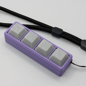 May include: A rectangular, lavender-colored keycap holder with four gray keycaps. A black lanyard is attached to the holder. The keycaps have a speckled appearance. The background is white.