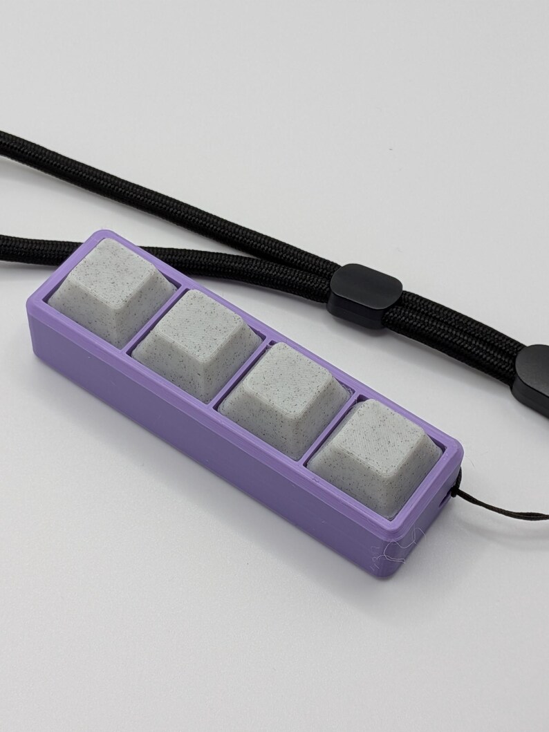 May include: A rectangular, lavender-colored keycap holder with four gray keycaps. A black lanyard is attached to the holder. The keycaps have a speckled appearance. The background is white.