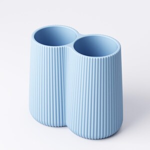 May include: A light blue, double-compartment container with vertical ribbed detailing. The two cylindrical openings are connected, creating a unique shape. The container is made of a solid, opaque material and sits on a white surface.