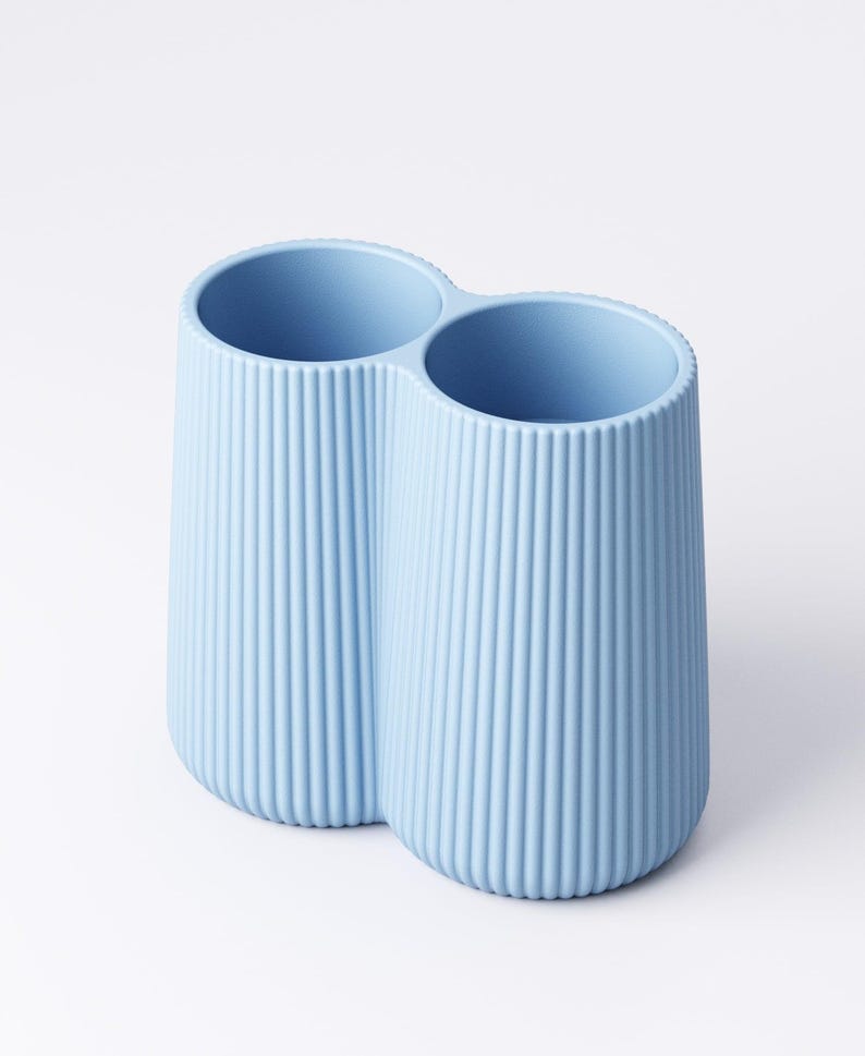 May include: A light blue, double-compartment container with vertical ribbed detailing. The two cylindrical openings are connected, creating a unique shape. The container is made of a solid, opaque material and sits on a white surface.