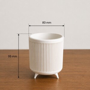 Puede incluir: Una maceta de cer&aacute;mica blanca con detalles acanalados verticales. La maceta cil&iacute;ndrica tiene tres patas peque&ntilde;as y mide aproximadamente 80 mm de di&aacute;metro y 95 mm de altura. El interior es liso y el exterior es texturizado.