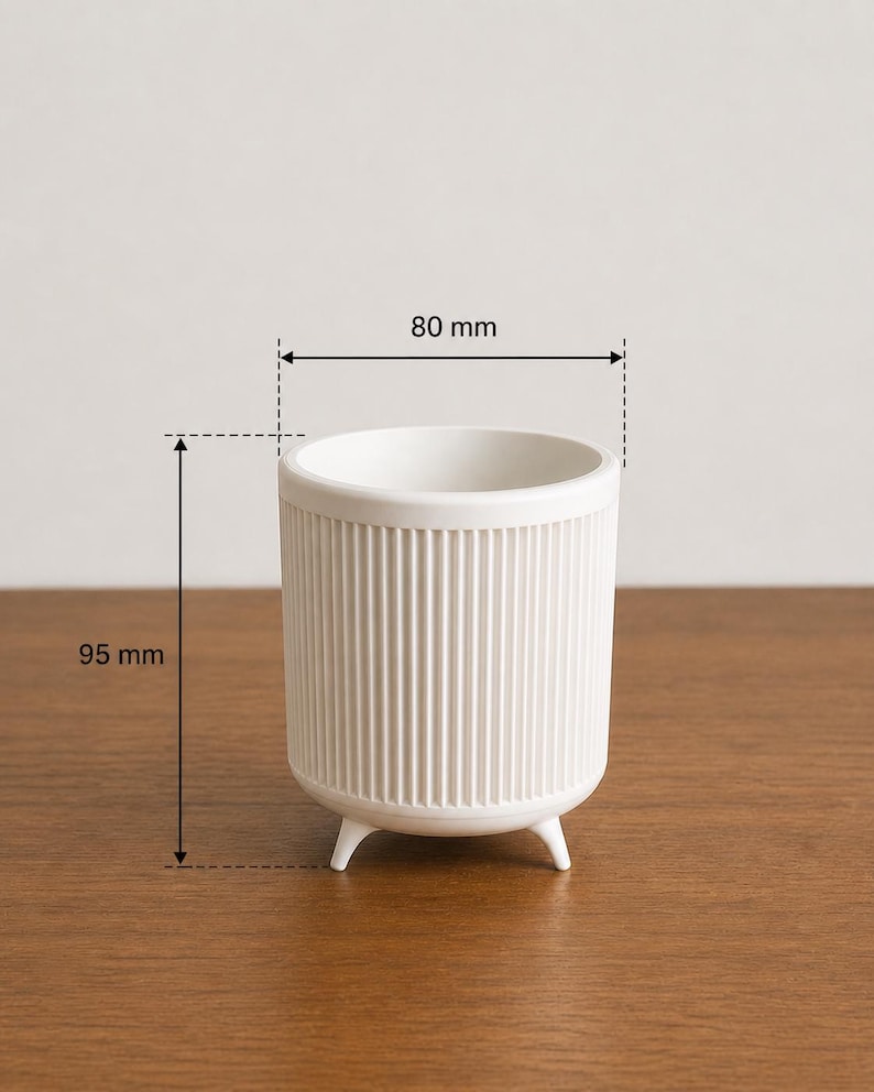 Puede incluir: Una maceta de cer&aacute;mica blanca con detalles acanalados verticales. La maceta cil&iacute;ndrica tiene tres patas peque&ntilde;as y mide aproximadamente 80 mm de di&aacute;metro y 95 mm de altura. El interior es liso y el exterior es texturizado.