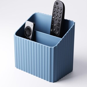 May include: A light blue, ribbed-textured remote control organizer. The organizer has two compartments and holds two black remote controls. The design is a solid, matte finish, and the container is made of a durable material.