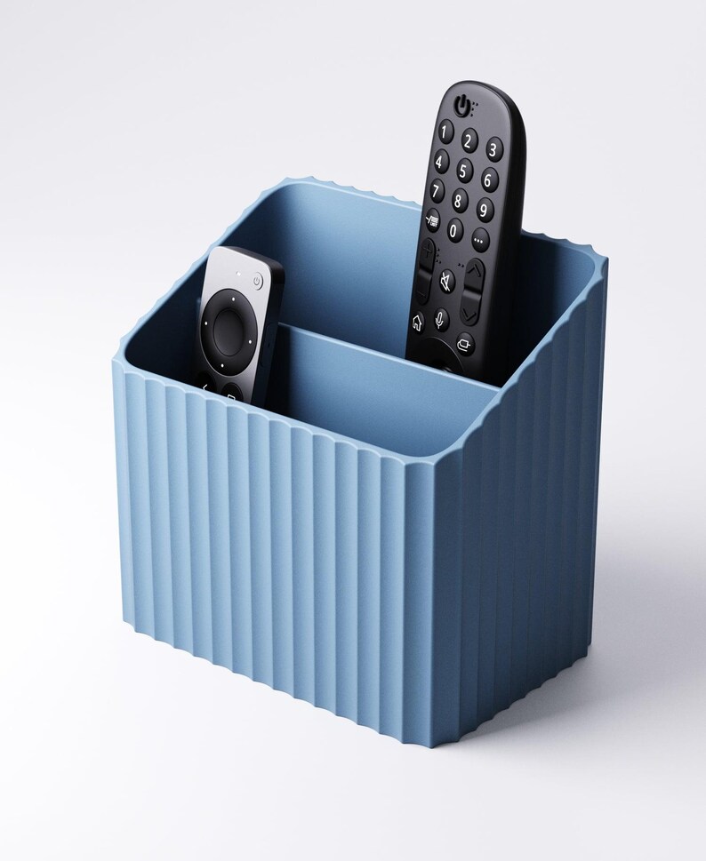 May include: A light blue, ribbed-textured remote control organizer. The organizer has two compartments and holds two black remote controls. The design is a solid, matte finish, and the container is made of a durable material.