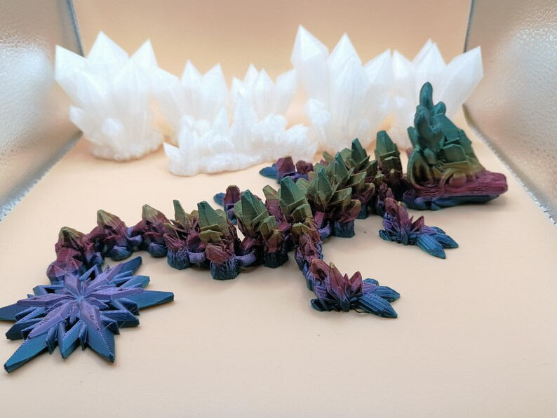 May include: A collection of 3D-printed crystal sculptures in various shapes and colors. The main piece is a multi-colored dragon-like figure with crystal formations. Additional white crystal clusters and a blue snowflake-shaped piece are also present.