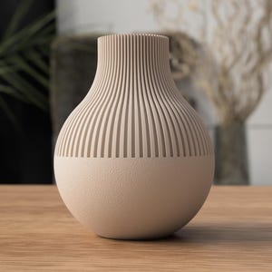 May include: A beige ceramic vase with a textured, ribbed upper section and a smooth, rounded base. The vase is displayed on a wooden surface, showcasing its neutral color and modern design. The vase is approximately 10 inches tall.