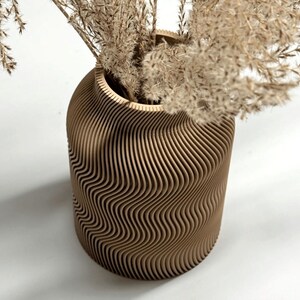 May include: A decorative vase with a unique, wavy design. The vase is made of light brown wood and is filled with dried, beige-colored plants. The vase has a cylindrical shape with a wide opening.