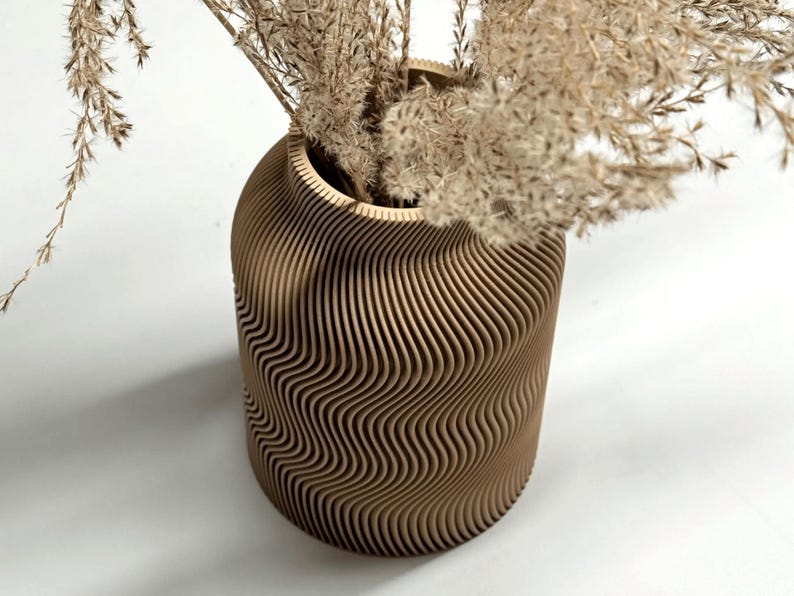 May include: A decorative vase with a unique, wavy design. The vase is made of light brown wood and is filled with dried, beige-colored plants. The vase has a cylindrical shape with a wide opening.