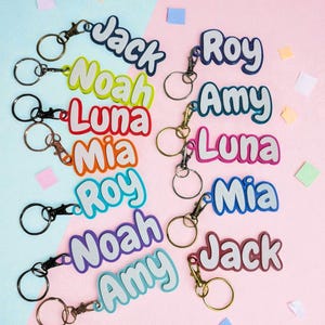 May include: A collection of colorful name keychains with various names like "Jack", "Noah", "Luna", "Mia", "Roy", and "Amy". Each keychain features a name in a bubble letter style with a contrasting outline and a metal clasp and ring.