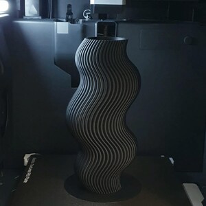 May include: A tall, wavy, black vase with a textured, ribbed surface. The vase has a wide base and a flared top. The design is modern and abstract, and the vase appears to be 3D-printed.