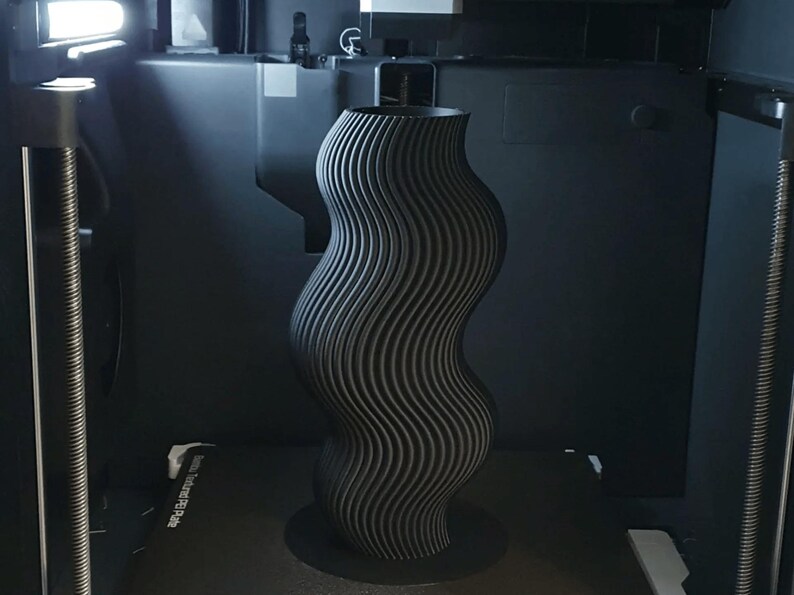 May include: A tall, wavy, black vase with a textured, ribbed surface. The vase has a wide base and a flared top. The design is modern and abstract, and the vase appears to be 3D-printed.