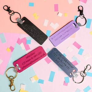 May include: Four rectangular keychains in black, purple, pink, and blue, each with a metal clasp and ring. The keychains display the name "John Doe", an address, and an email address. The background is decorated with colorful confetti.