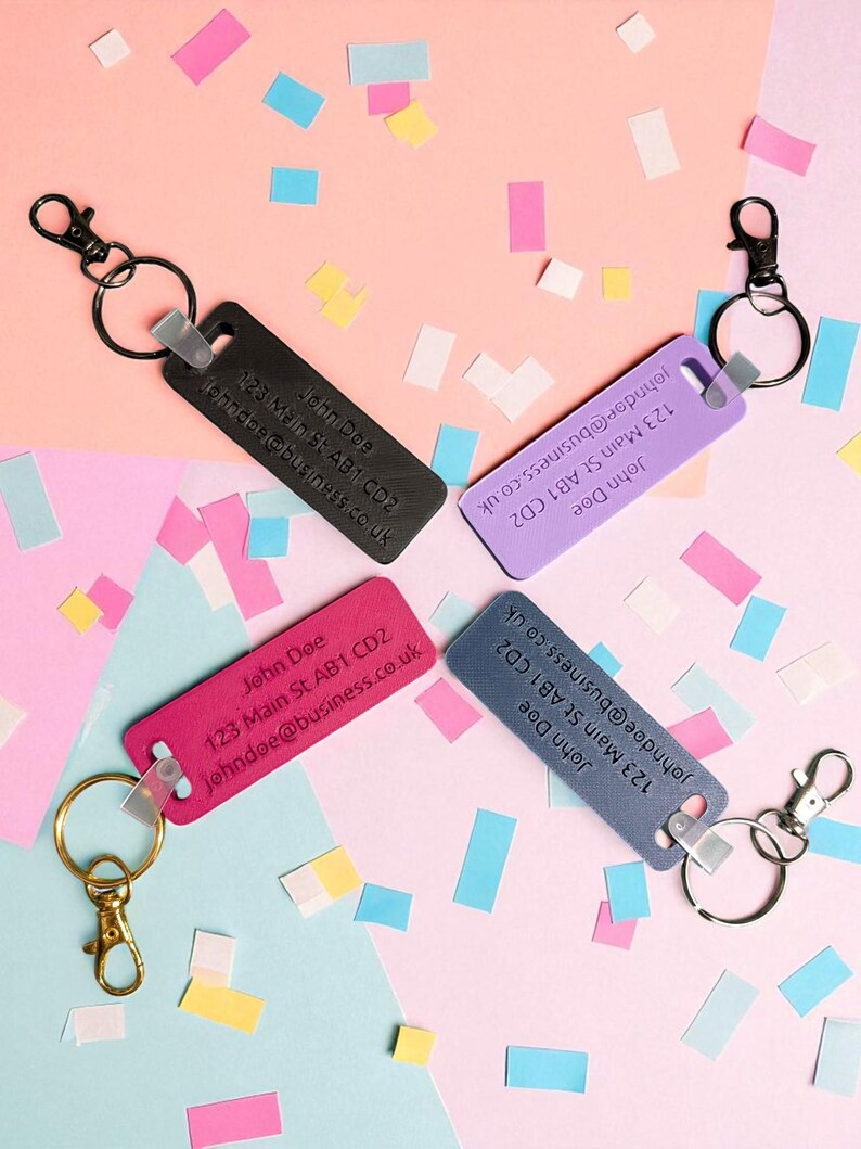 May include: Four rectangular keychains in black, purple, pink, and blue, each with a metal clasp and ring. The keychains display the name "John Doe", an address, and an email address. The background is decorated with colorful confetti.