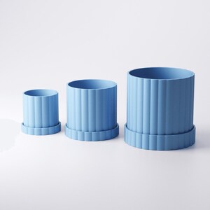 May include: Three light blue cylindrical planters of varying sizes, each with vertical ribbed detailing and a matching base. The planters are arranged from smallest to largest, showcasing a minimalist design.
