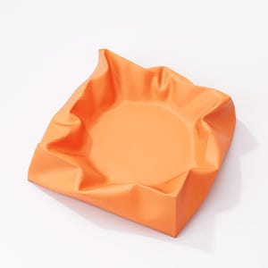 May include: An orange, square-shaped bowl with a slightly crumpled, folded rim. The bowl has a smooth, matte finish and sits on a white surface. The interior of the bowl is a lighter shade of orange.
