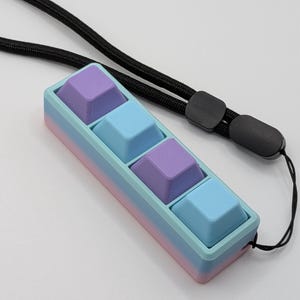 May include: A pastel-colored keyboard keycap holder with four square keycaps in shades of purple and blue. The holder is a gradient of pink, blue, and purple. A black lanyard is attached to the holder.