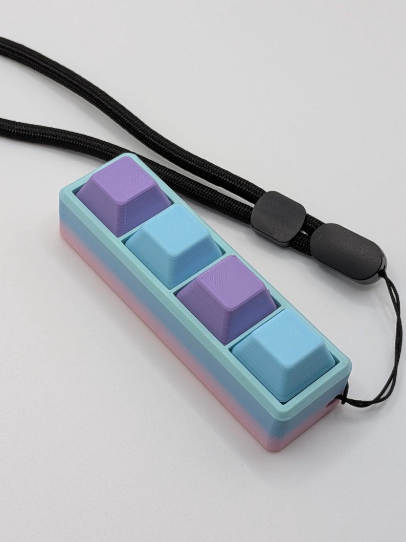 May include: A pastel-colored keyboard keycap holder with four square keycaps in shades of purple and blue. The holder is a gradient of pink, blue, and purple. A black lanyard is attached to the holder.