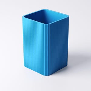 May include: A bright blue, rectangular pencil holder with vertical ridged detailing. The container has a square opening and is made of a solid, opaque material. The design is minimalist and modern, suitable for a desk or workspace.