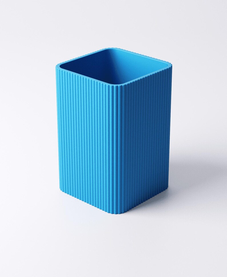 May include: A bright blue, rectangular pencil holder with vertical ridged detailing. The container has a square opening and is made of a solid, opaque material. The design is minimalist and modern, suitable for a desk or workspace.