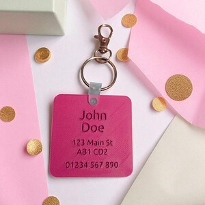 May include: A fuchsia square keychain with engraved text reading "John Doe", address, and phone number. The keychain has a metal clasp and ring. Gold confetti and pink paper are in the background.