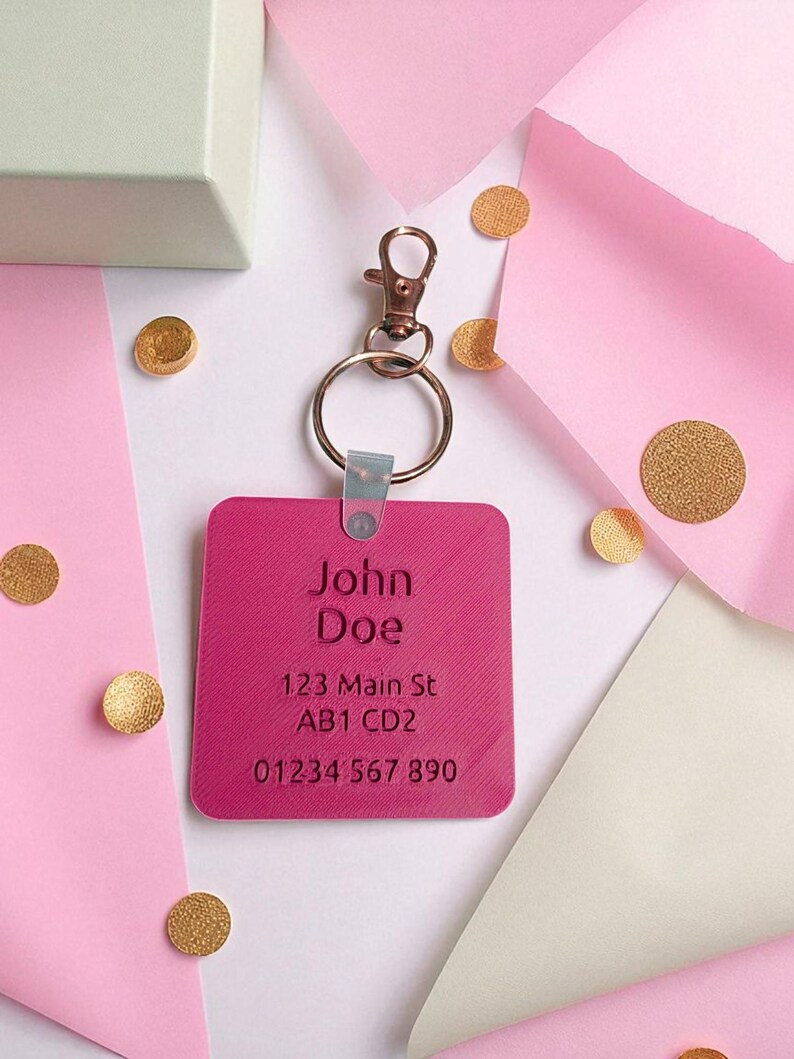 May include: A fuchsia square keychain with engraved text reading "John Doe", address, and phone number. The keychain has a metal clasp and ring. Gold confetti and pink paper are in the background.