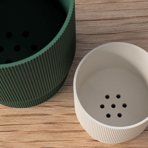 May include: Two cylindrical containers with ribbed sides and a series of holes in the bottom. One is dark green, the other is off-white. The containers are on a light brown wooden surface.