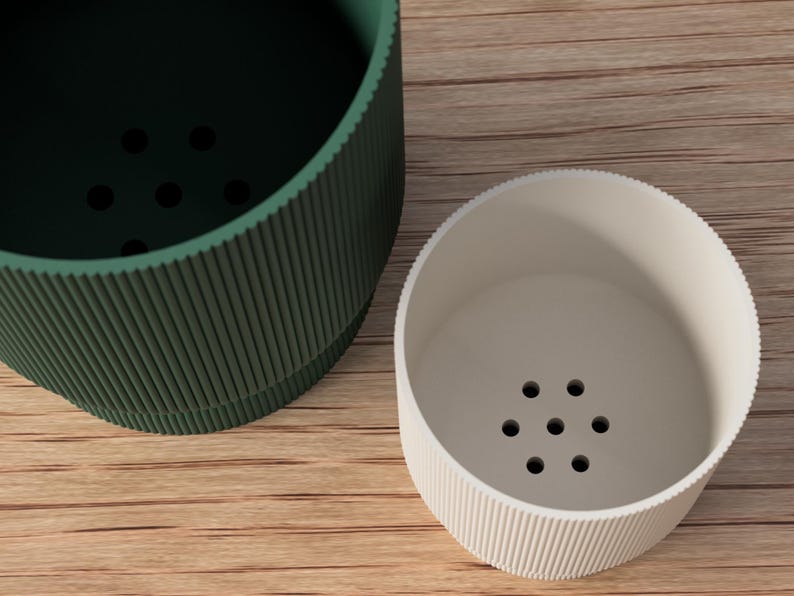 May include: Two cylindrical containers with ribbed sides and a series of holes in the bottom. One is dark green, the other is off-white. The containers are on a light brown wooden surface.