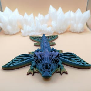 May include: A blue and purple dragon figurine with intricate wing details and a crystal formation in the background. The dragon has a detailed head and body, with a gradient of colors. The crystals are white and have a jagged appearance.