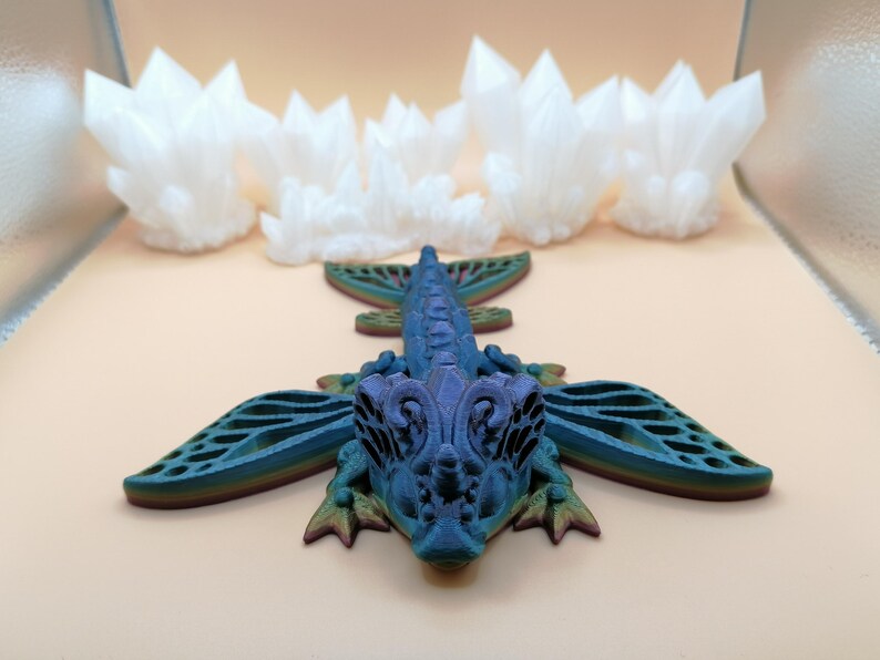 May include: A blue and purple dragon figurine with intricate wing details and a crystal formation in the background. The dragon has a detailed head and body, with a gradient of colors. The crystals are white and have a jagged appearance.
