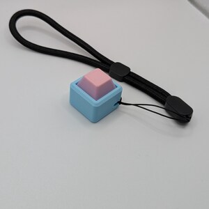 May include: A small, square, blue and pink accessory with a black wrist strap. The accessory has a light blue square base with a pink and purple pyramid-shaped top. The black wrist strap is made of woven material.