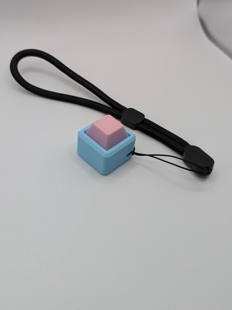 May include: A small, square, blue and pink accessory with a black wrist strap. The accessory has a light blue square base with a pink and purple pyramid-shaped top. The black wrist strap is made of woven material.