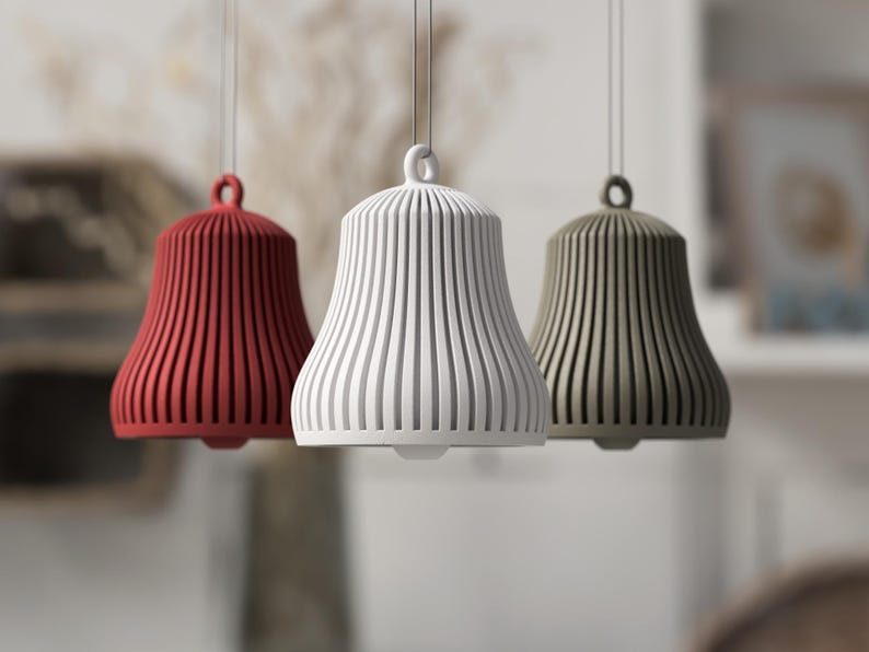 May include: Three pendant lights in red, white, and olive green. Each bell-shaped light has vertical grooves and a loop at the top for hanging. The lights are suspended from thin cords against a blurred background.